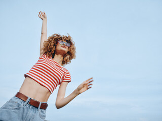 A joyful young woman with curly hair in a red striped crop top and jeans spins against a vast blue...