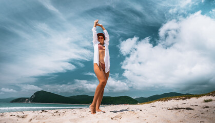 Confident woman stretching arms up while standing on warm sandy beach. Wearing bikini, white shirt and sunglasses, she enjoys summer breeze and freedom of movement under tropical sky.