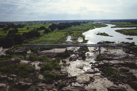 Aerial view of a bridge crossing a rugged, rocky riverbed amidst lush green vegetation, edged by trees under a vast sky, Kano, Kano, Nigeria.