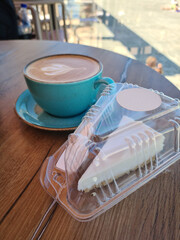 A turquoise coffee cup sits on a wooden table next to a slice of cake in a clear plastic container....