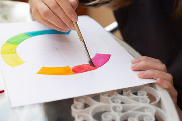 Close-up of a brush applying red watercolor to a segment of a color wheel. White paper on a...