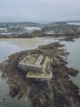 Aerial view of the National Fort standing firm against the relentless sea, with the distant city offering a stark contrast of stone and sky, Saint-Malo, Brittany, France.