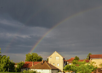 Double rainbow over gray sky with sunlit houses and trees below