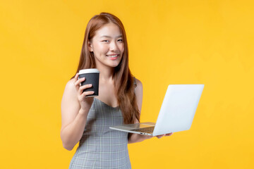 Smiling young Asian woman holding laptop computer and takeaway coffee cup with cheerful confident expression, working and studying online on yellow background, technology and lifestyle concept