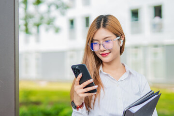 Fototapeta premium Asian female student in warm clothes using phone to chat with friends and ready for video call while in university.