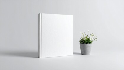 Fototapeta premium Blank white hardcover book mockup standing next to a small plant in a concrete pot.