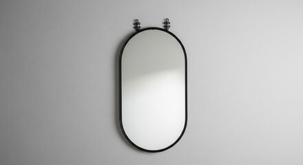 Obraz premium A sleek, modern oval mirror with a black metal frame hangs on a plain white wall. The minimalist design is complemented by two small, decorative knobs or hangers at the top, adding a unique