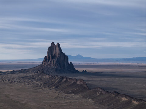 Aerial view of Shiprock's jagged, dark volcanic rock formation rises dramatically from the flat, arid desert landscape under a vast sky, Shiprock, New Mexico, United States.