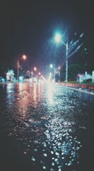 A blurry night scene depicts a wet asphalt road reflecting streetlights. Rain falls in soft focus, creating an ethereal glow and muted color palette