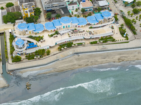 Aerial view of a coastal promenade with bright blue rooftops contrasting against the beige sand and the turquoise waves of the ocean, Puerto Colombia, Atlantico, Colombia.