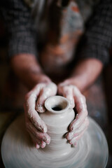 Caucasian middle aged man shaping clay vase on pottery wheel, hands covered in wet clay, focusing...