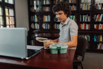 Student studying in library with laptop and book, enjoying a coffee break