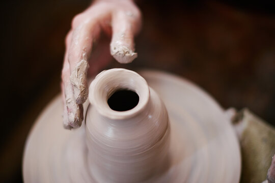 Caucasian adult hand shaping clay on pottery wheel, fingers covered in wet clay, creating ceramic vessel during pottery making process, close up of skilled craftsmanship in action