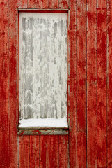 A striking red wooden wall features an empty window frame, with a dusting of snow at its base, conveying a cold winter atmosphere.