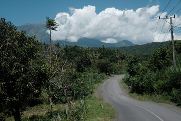 Curving road through a lush tropical forest with mountain backdrop and dramatic white clouds.