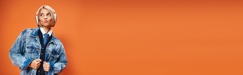 Blonde young woman poses stylishly in trendy denim jacket against bright orange backdrop