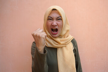 Angry Young Asian Woman Clenching One Fist and Shouting Against Pink Background.