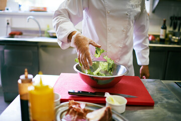 Midsection of man wearing chef uniform preparing salad in commercial kitchen, hand tossing fresh...