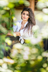 Stylish  brunette sensual woman with white shirt and denim  in nature, leaning on wooden fence near...