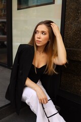 A young woman sits casually on a ledge, wearing stylish attire that combines a black top and white...