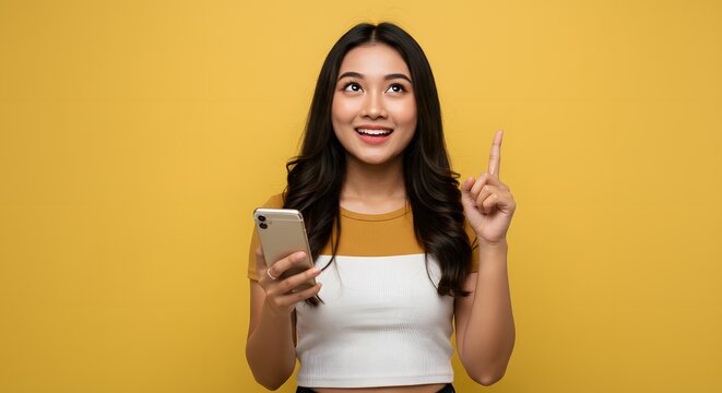 Excited Asian Woman Holding Phone Pointing Upwards, Yellow Background