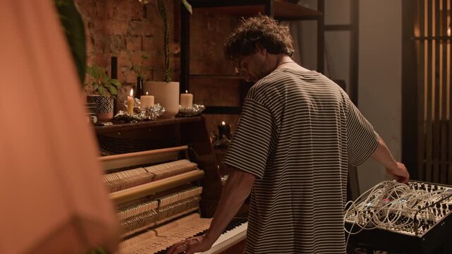 Rear view of middle aged male musician playing piano adding electronic sound design to classical music by tweaking modular synth while live performing in room decorated with candles