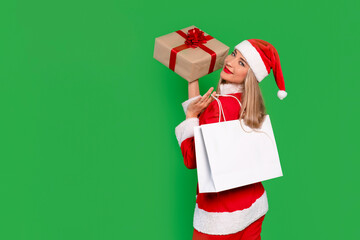 Joyful woman dressed as Santa Claus holding a gift and shopping bag against a bright green background during the holiday season