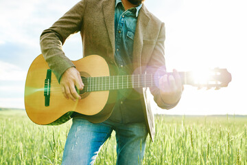 Caucasian young adult man playing acoustic guitar outdoors in field, strumming instrument with both...