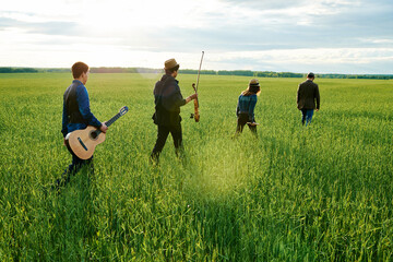 Group of young adult multiethnic musicians walking through green field carrying guitar and violin,...