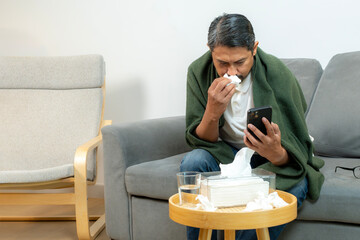 A young man with fever and runny nose blowing into tissue and taking medicine while resting at...