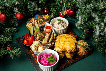 Festive holiday food assortment on black board with Christmas tree background. Includes salads, pickles, mushrooms, caviar, nuts, and baked dishes. Traditional celebration setup