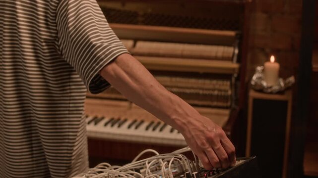 Middle aged male musician playing piano then altering sound in modular synthesizer while experimenting with sound design for new music album in recording studio decorated with candles