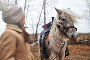 Caucasian child girl standing outdoors facing gray horse with saddle, autumn forest background...