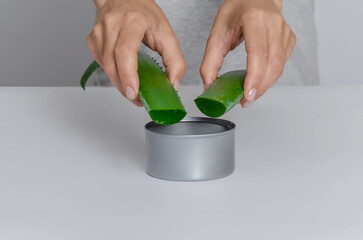 Unrecognizable woman holding two freshly cut aloe leaves above jar of gel on white table, front view