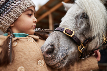 Caucasian child girl wearing knit hat hugging small pony outdoors, showing gentle interaction...