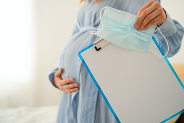 Future mother in medical mask holds pristine notebook awaiting important birth notes. Sterile yet warm hospital environment for preparation.