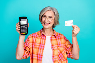 Senior woman holding a card reader and credit card against a bright background showing joy and...
