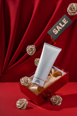 White bottle cosmetic product in red giftbox levitate surrounded by layered gold roses on red background. Mock up design for advertising, seasonal discounts. Valentine's Day banner