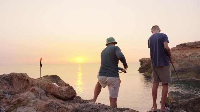 Silhouetted parent fishing with child during golden sunrise, sharing tranquil moment on rocky coastal shoreline with soft morning light reflecting across ocean surface