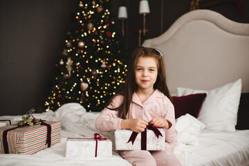 Smiling kid girl 4-5 year old wearing pajamas open Christmas box presents sitting in bed over xmas...