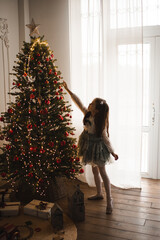 Stylish child girl 4-5 year old wearing princess dress decorating Christmas tree in living room at...