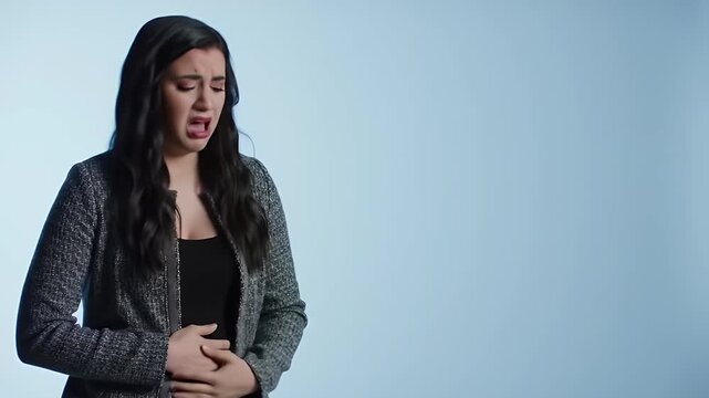 Young Woman in Black Top with Dark Hair Experiencing Abdominal Pain Against Light Blue Background Under Bright Studio Lights Expressing Discomfort Holding Stomach in Distress Medical Concept
