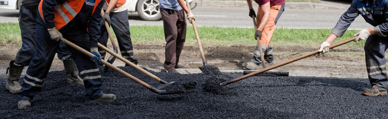 Road crew with shovels spreads and levels fresh asphalt on a city street; hot mix, hi-vis vests and...