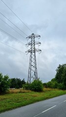 Energy communication pole on the street, sky. A tall lattice steel transmission tower carrying several power lines stands against a background of trees bushes and blue sky