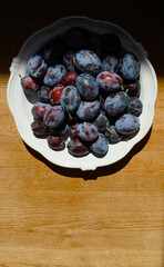 Close-up of fresh plums in a white ceramic bowl on a wooden surface. Summer harvest illuminated by soft natural light