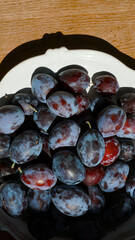 Close-up of fresh plums in a white ceramic bowl on a wooden surface. Summer harvest illuminated by...