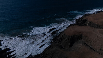 Rocky coastline with powerful waves crashing against rugged cliffs under a clear sky, showcasing the contrast between deep blue ocean and barren land.