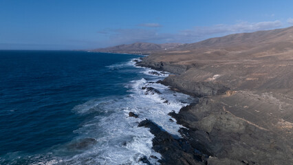 Rocky coastline with powerful waves crashing against rugged cliffs under a clear sky, showcasing the contrast between deep blue ocean and barren land.