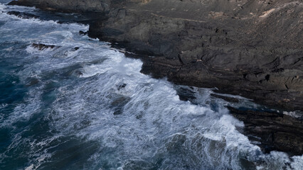Rocky coastline with powerful waves crashing against rugged cliffs under a clear sky, showcasing the contrast between deep blue ocean and barren land.
