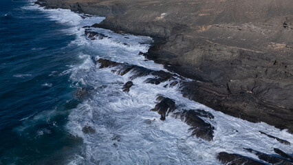 Rocky coastline with powerful waves crashing against rugged cliffs under a clear sky, showcasing the contrast between deep blue ocean and barren land.